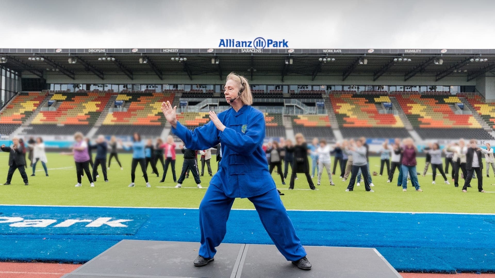 Chris Jones, in a blue Taiji uniform with a headset microphone, leads a large group of older people through Taiji on the pitch at Saracens' Allianz Park, under grey skies, at an Age UK Barnet event in 2016