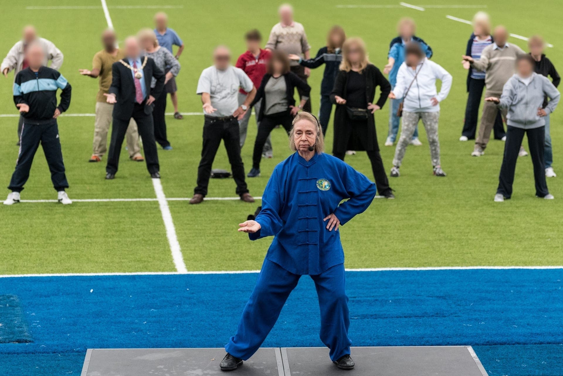 Chris Jones in blue leads Taiji from a low stage on the pitch at Allianz Park, with rows of older people following behind her, among them the Mayor of Barnet in a suit and chain of office