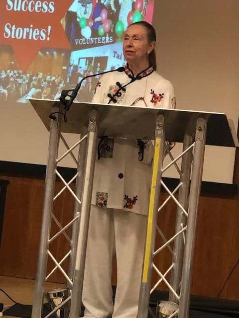 Chris Jones in a white Taiji suit embroidered with flowers stands at a lectern in front of a Success Stories slide at the Kensington & Chelsea Forum AGM