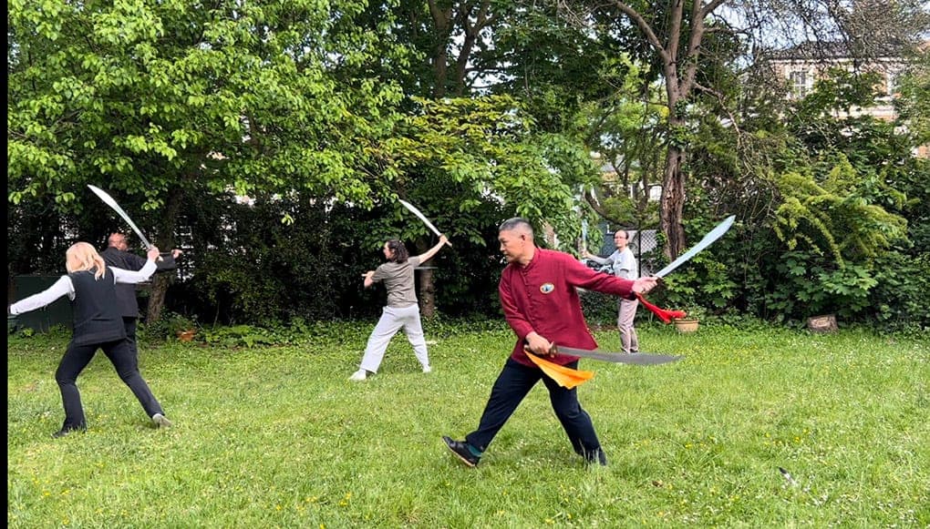 A teacher in a red jacket leading students through a double-sabre routine on a lawn among trees