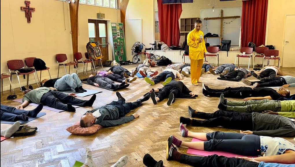 Students lying on mats in a hall for a guided relaxation, a teacher in yellow standing among them