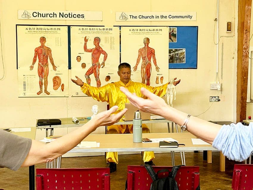 A teacher in yellow silk leading a Qigong health workshop in front of acupuncture charts, two students' outstretched arms in the foreground