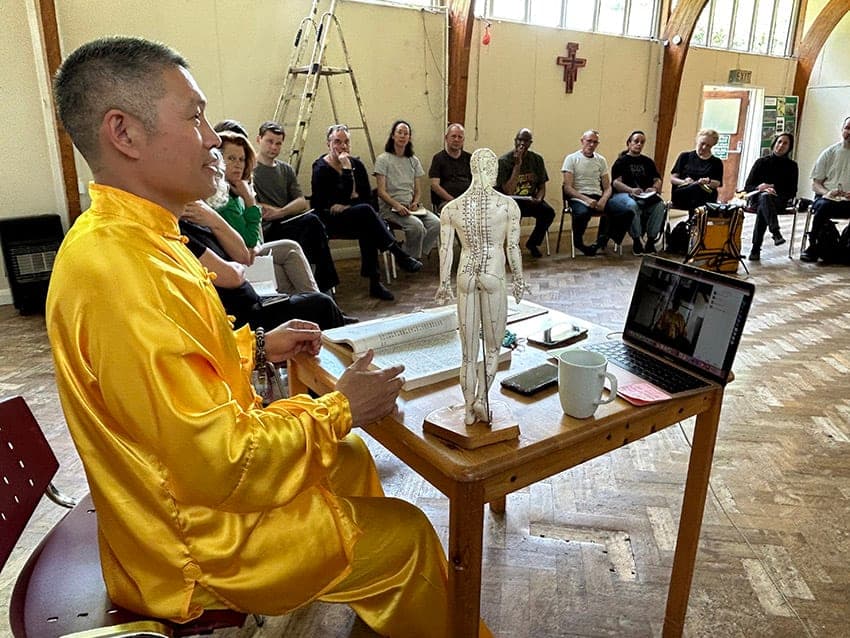 A teacher in yellow silk teaching at a table with an acupuncture model and a laptop, students seated behind