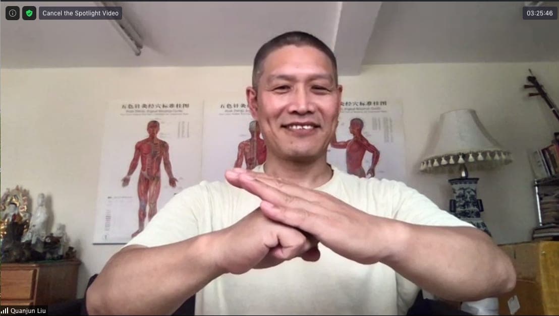 Shifu Liu Quanjun smiling and giving the martial salute to the camera during an online class, acupuncture charts on the wall behind him