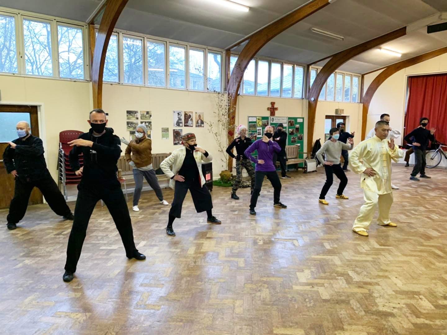 Shifu Liu Quanjun, in pale yellow silk, leads students, most in face masks, through Chen Foundation training in a church hall with arched wooden beams