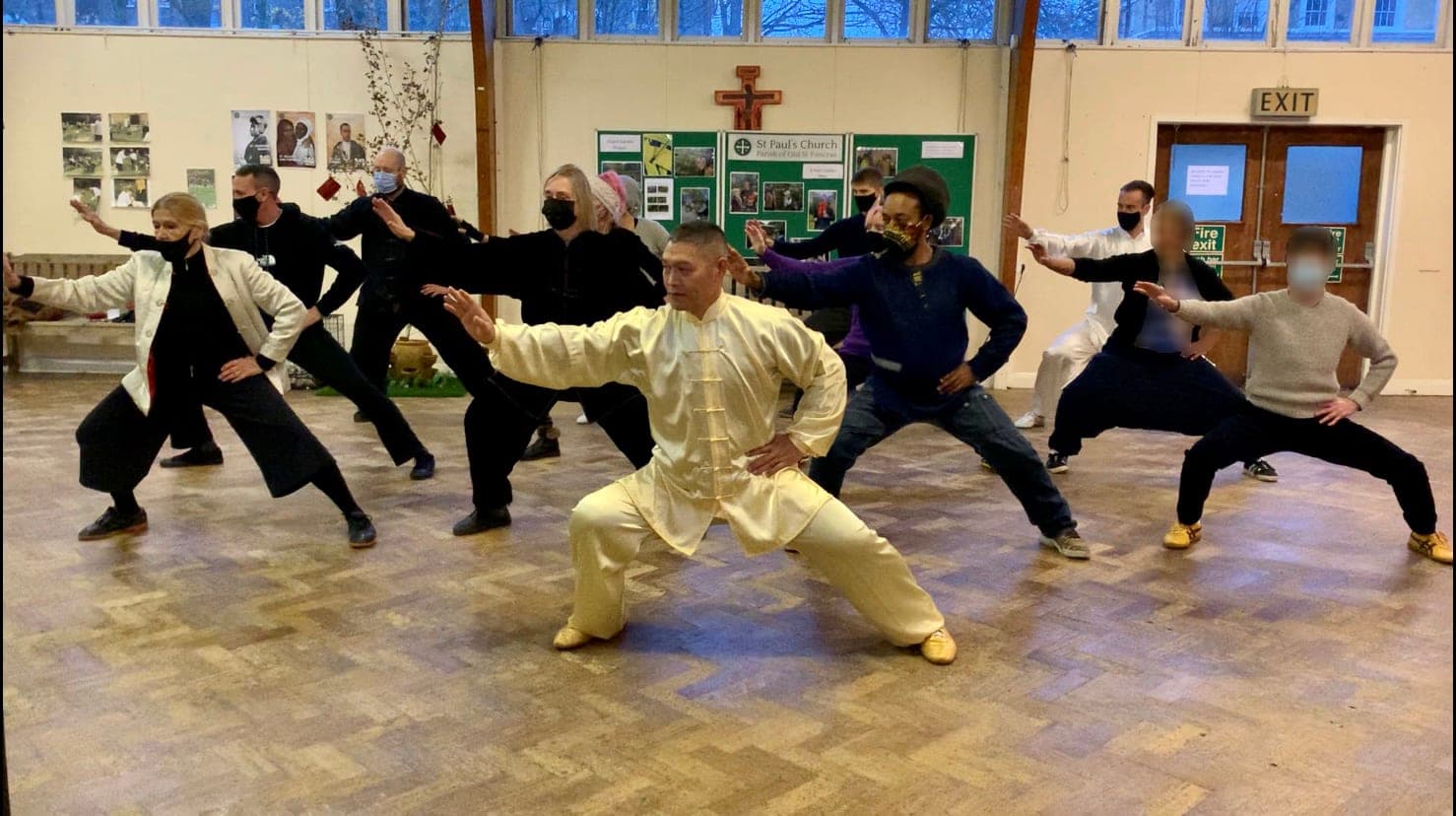 Shifu Liu Quanjun in pale yellow silk in a low stance at the front of St Paul's Church hall, with masked students following the movement behind him