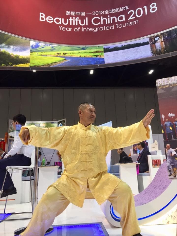 Shifu Liu Quanjun in yellow silk holding a wide Taiji stance with arms outstretched, beneath a Beautiful China 2018 banner