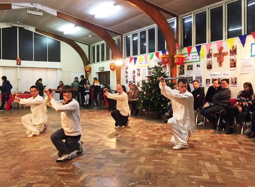 Four students kneel low with swords raised during a sword form demonstration beside a Christmas tree