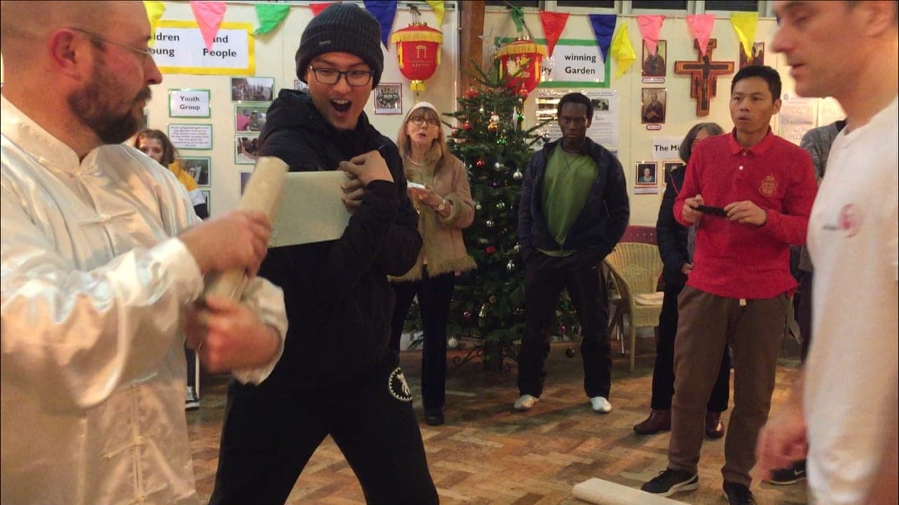 Gary, in white silk, holds up the two halves of a broken concrete block while onlookers react beside a Christmas tree