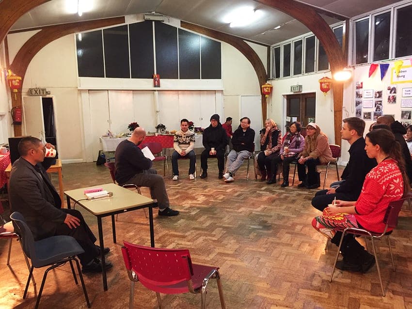 Guests sit in a circle of chairs listening to a storyteller reading at a small table