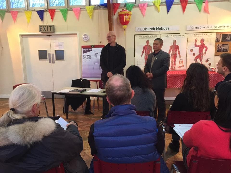 Torben Hersberg and Shifu Liu stand before a seated audience in a hall hung with bunting and acupuncture charts