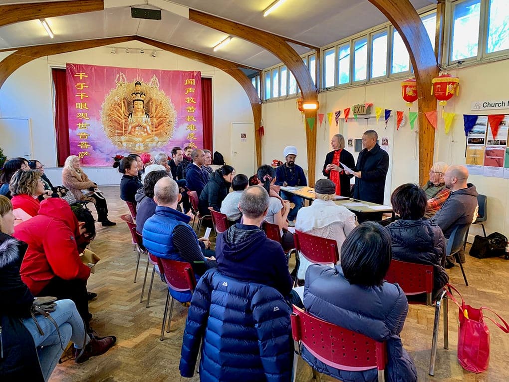 Shifu Liu stands beside a presenter reading from a sheet as the 2019 awards are announced to a seated audience in front of a pink Guanyin banner