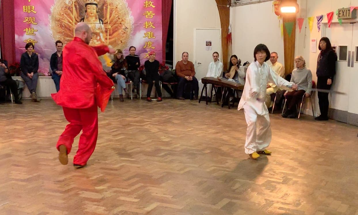 Two performers, one in red silk and one in white, circle each other with sabres on a parquet floor while an audience watches