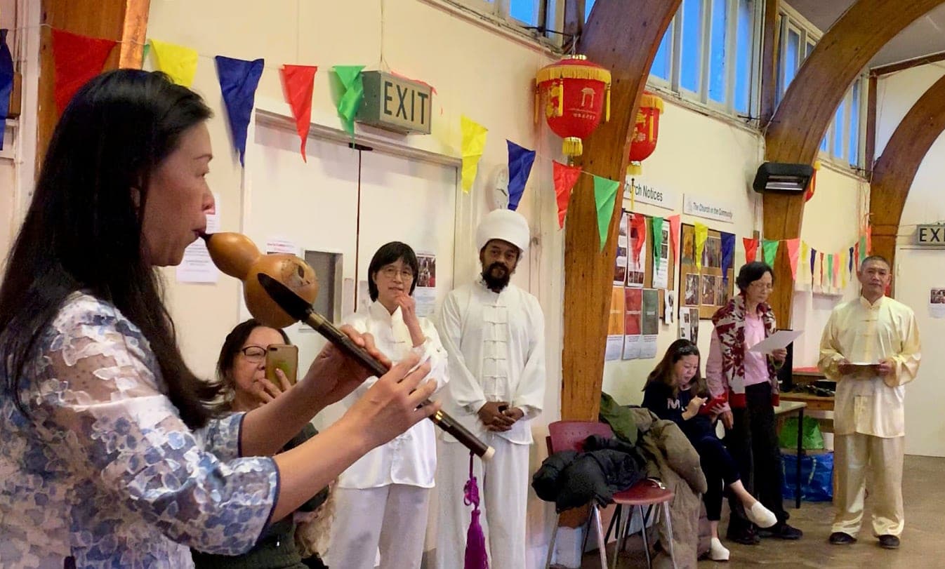 Joanna Zhenghui Qiu plays a gourd flute while students in white and Shifu Liu stand listening