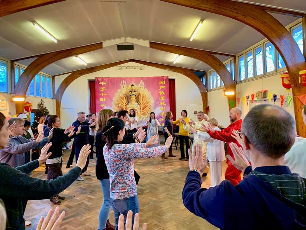 A large circle of students and guests follow a Qigong movement with raised palms in a church hall with wooden arches and a pink Guanyin banner