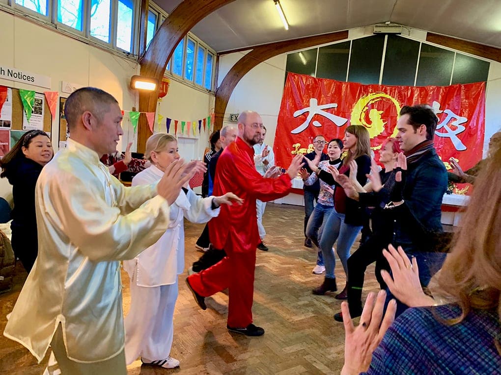 Shifu Liu in cream silk leads guests through a Qigong movement in front of the red Taiji banner