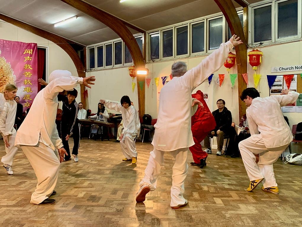 A group in white silk performs a Xinjia form with arms outstretched on the parquet floor
