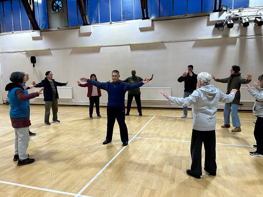 A teacher and a circle of students practising standing Qigong with arms spread wide in a sports hall