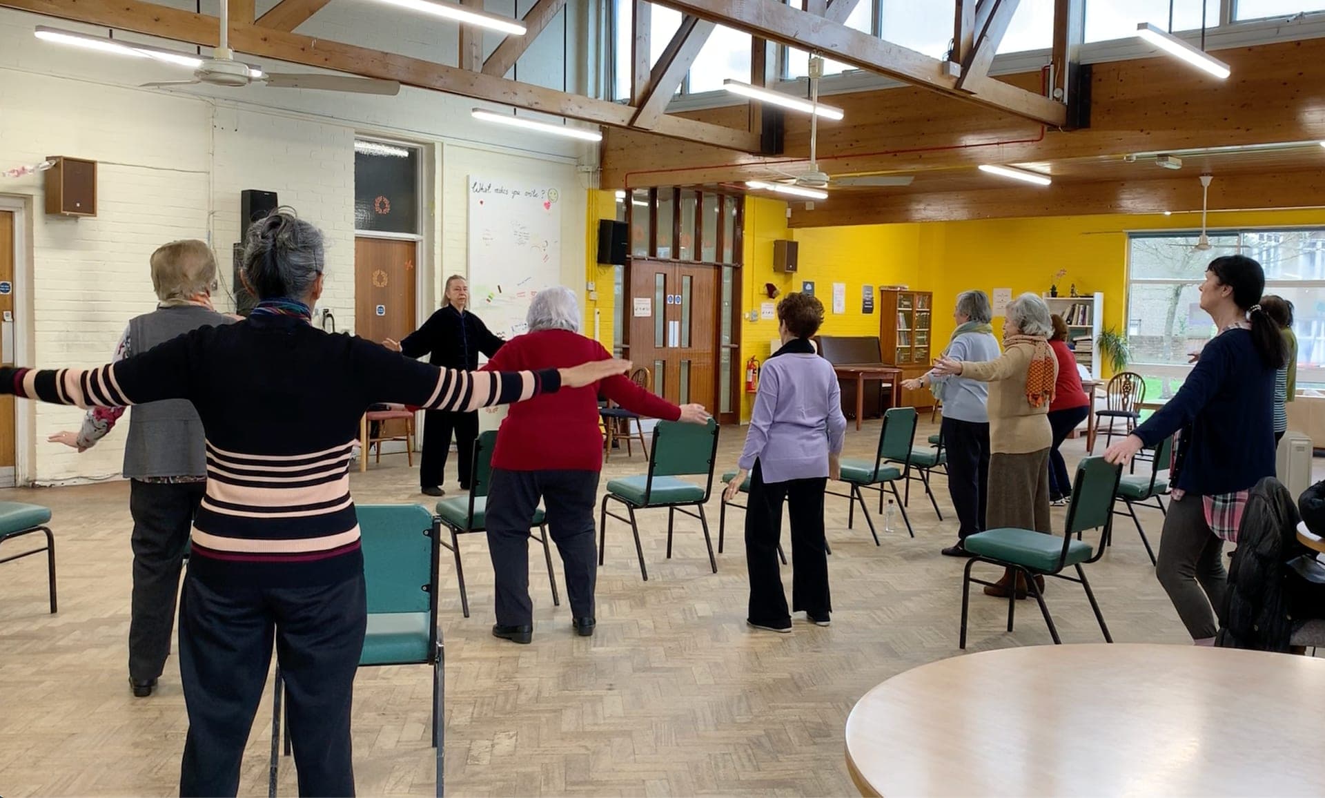 Chris Jones in black teaches a class of older people standing between chairs in a bright community hall with a yellow wall, 2020