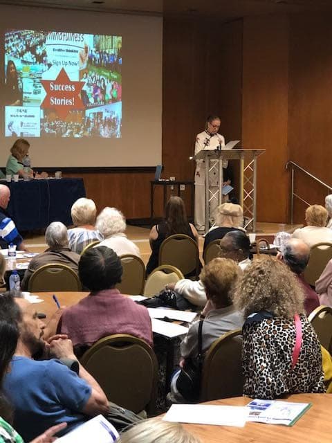 Chris Jones in a pale Taiji jacket speaks at a lectern to a seated audience of older people, with a slide about mindfulness projected behind her