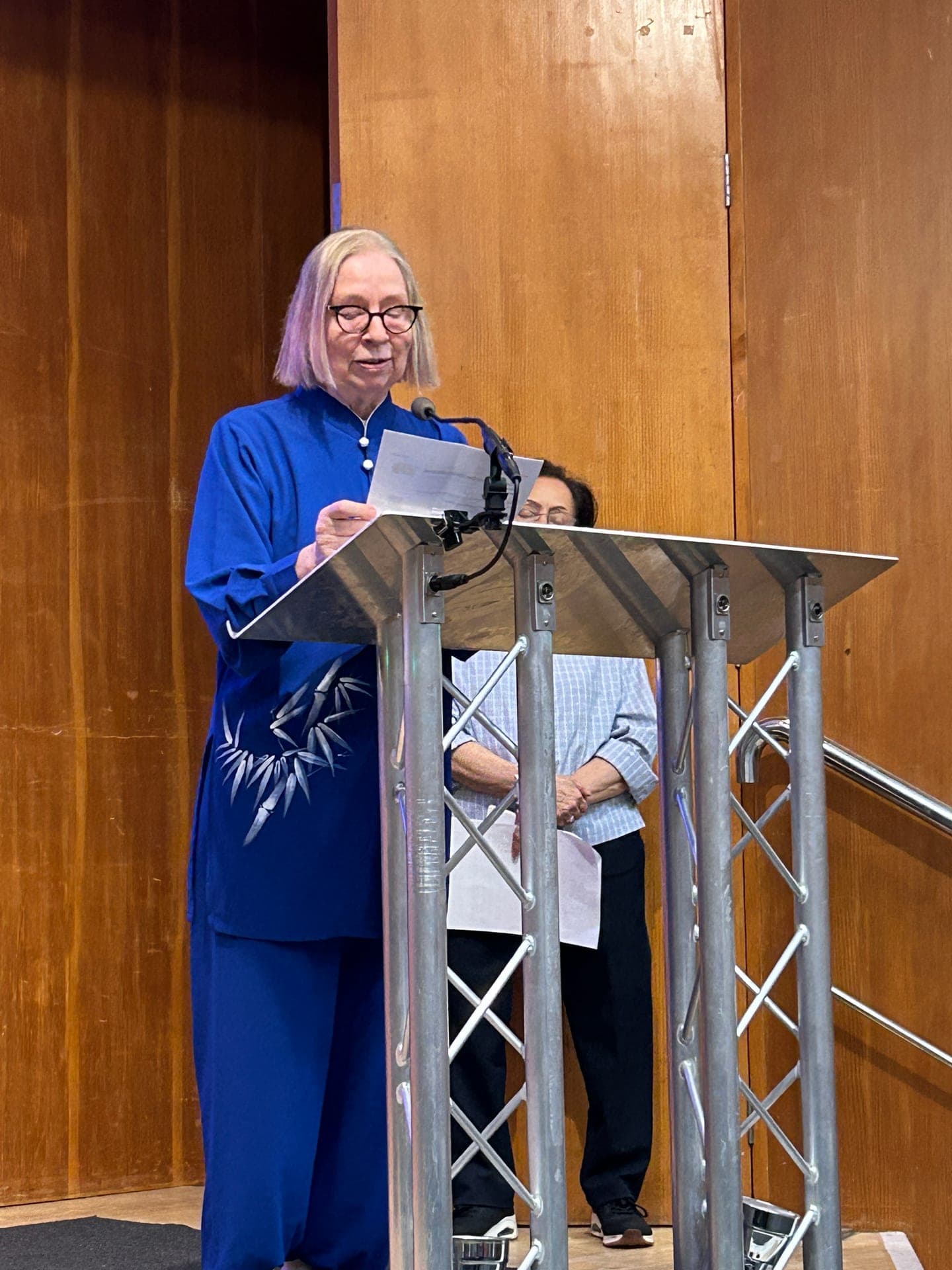 Chris Jones in a blue bamboo-patterned Taiji suit reads from notes at a lectern at the Kensington & Chelsea Forum AGM, 2025