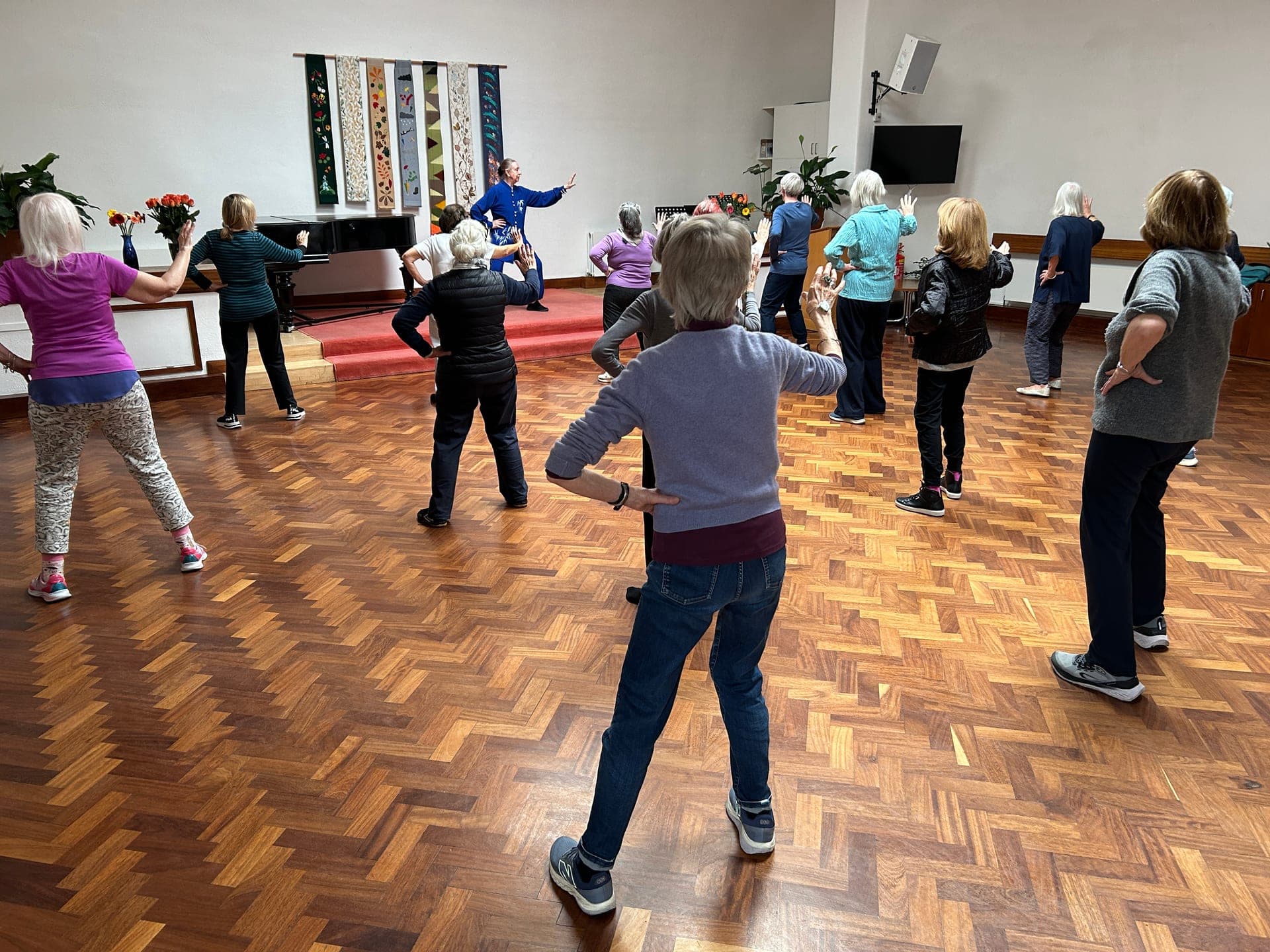 Chris Jones in blue leads the Kensington & Chelsea Forum class from the front of a hall with a grand piano and hanging textile banners, October 2025
