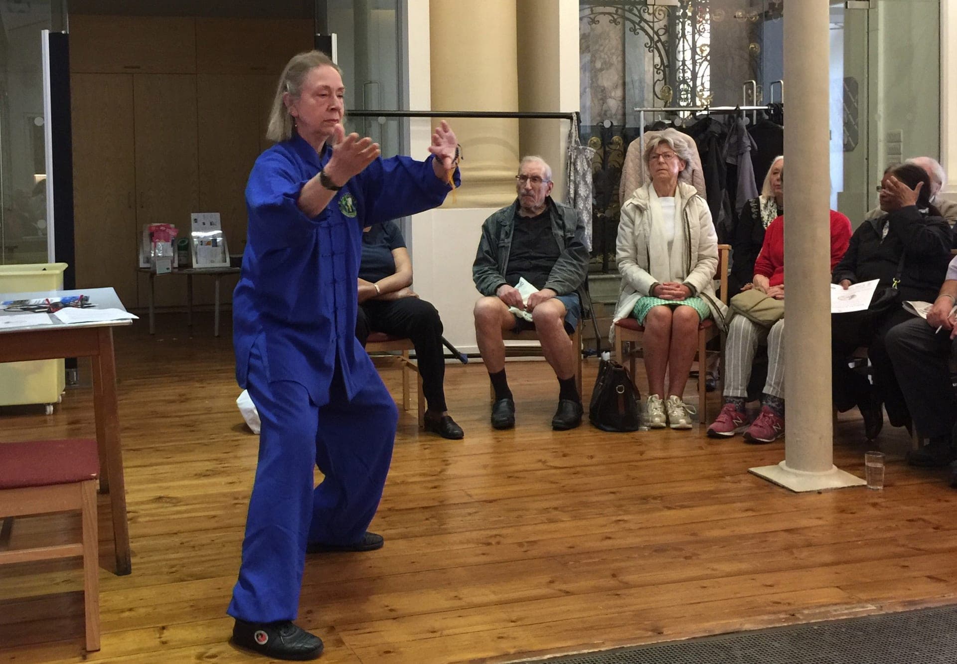 Chris Jones in blue leads a room of older people, some seated and some standing, with arms extended forward, at a Kensington & Chelsea Forum talk in 2018