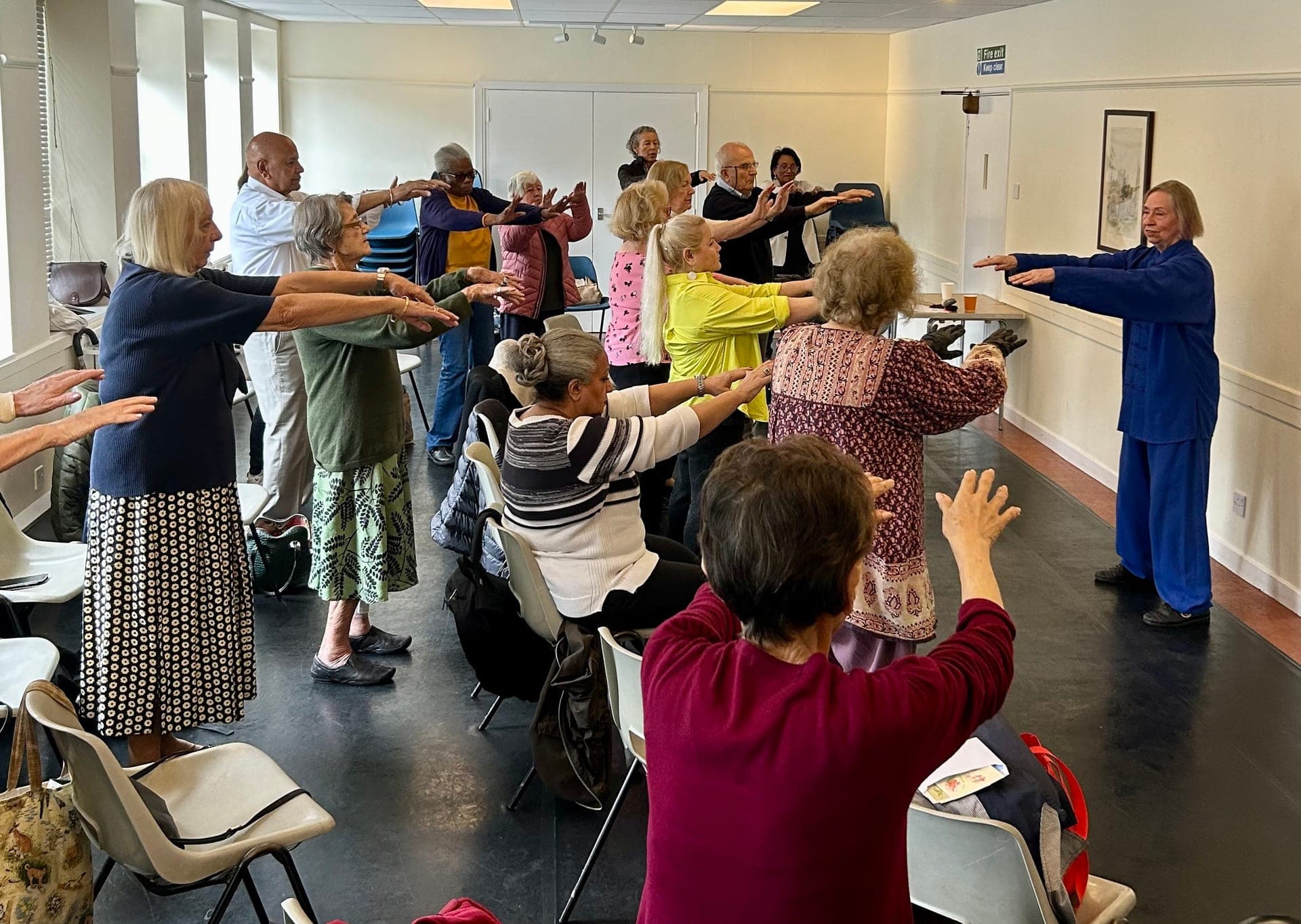 Chris Jones in blue demonstrates a Taiji posture on a wooden floor to a row of seated older people, 2023