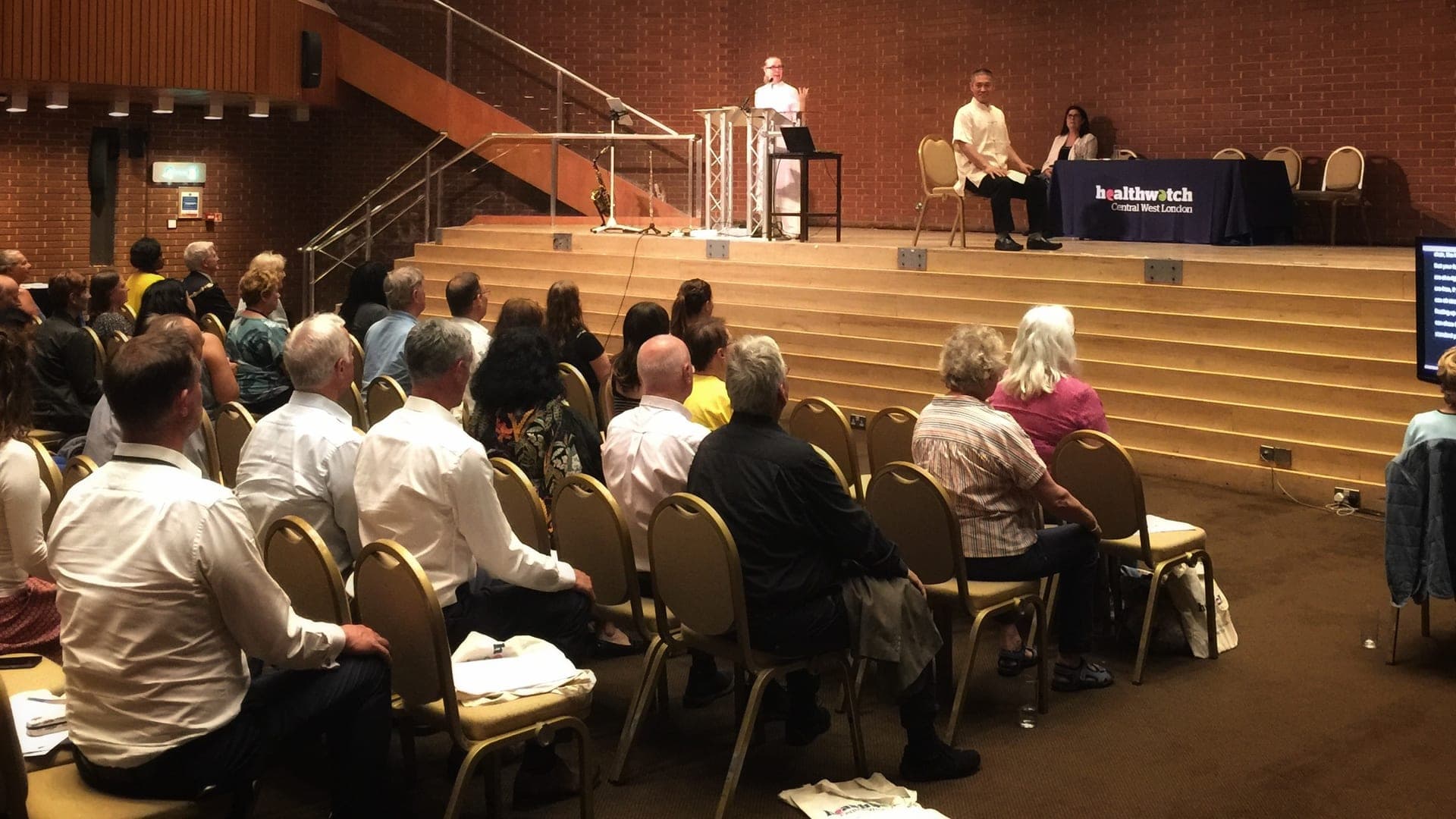 Chris Jones speaks from a lectern on the stage of the brick-walled hall at Kensington Town Hall, Shifu Liu seated beside a Healthwatch Central West London table, with the seated audience in the foreground