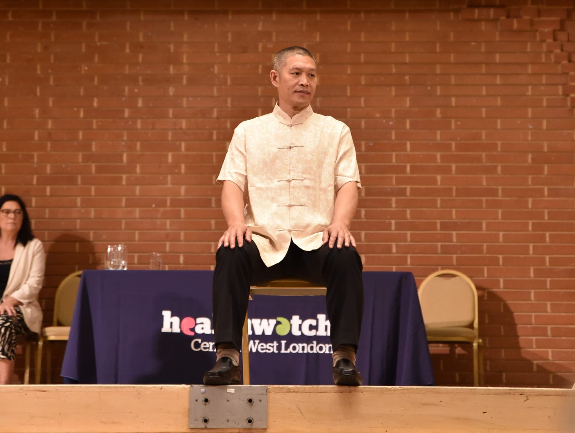 Shifu Liu Quanjun in a cream silk shirt sits upright on a chair at the front of the stage, hands on his knees, in front of a Healthwatch Central West London banner