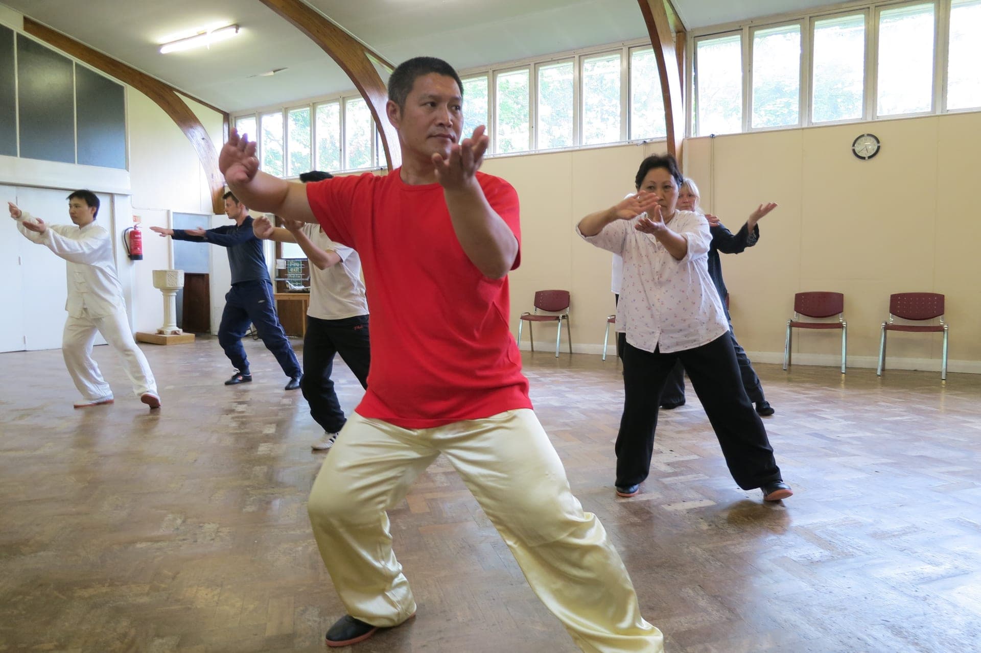 A silk reeling class in a light hall with arched timber beams, a man in a red T-shirt in front turning both hands outward as others practise behind him