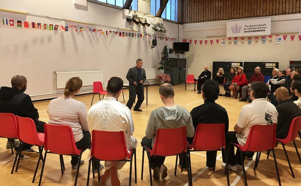 Shifu Liu, in a dark suit, demonstrates in the middle of a sports hall to students seated in a circle of red chairs