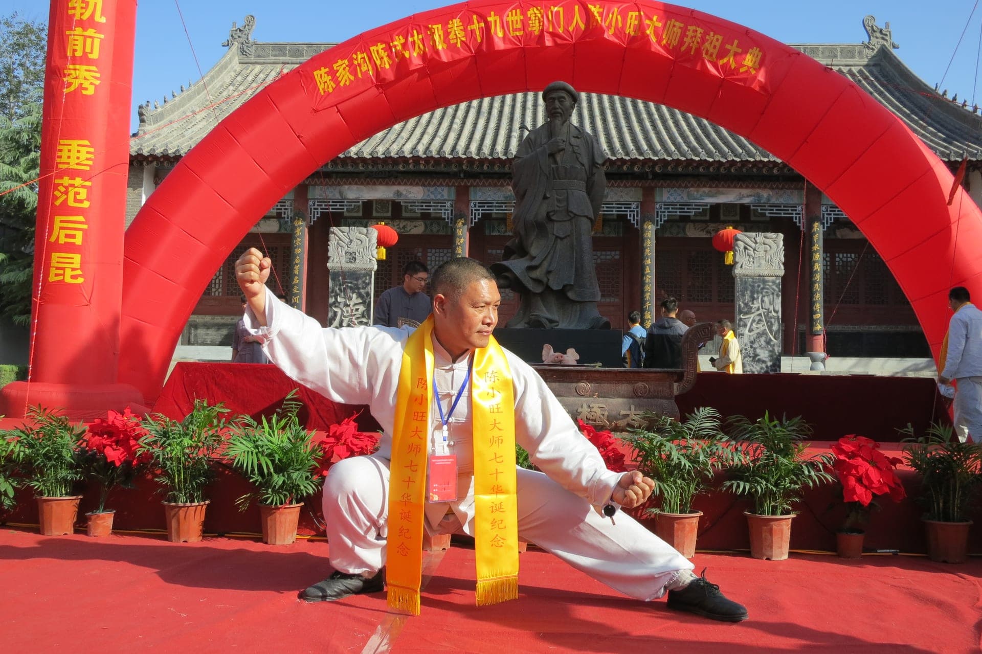 Shifu Liu Quanjun in a low stance on a red stage beneath a red arch, in front of the Chen family ancestral temple in Chenjiagou