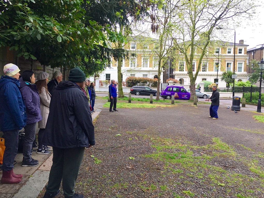 Shifu Liu demonstrates the form on a gravel path while a group of people watches from the side of the square