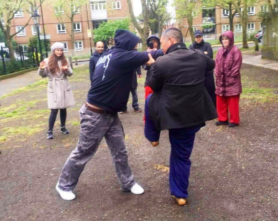 Shifu Liu in a dark coat demonstrates a martial application with a student in a hooded top, while others watch in the square
